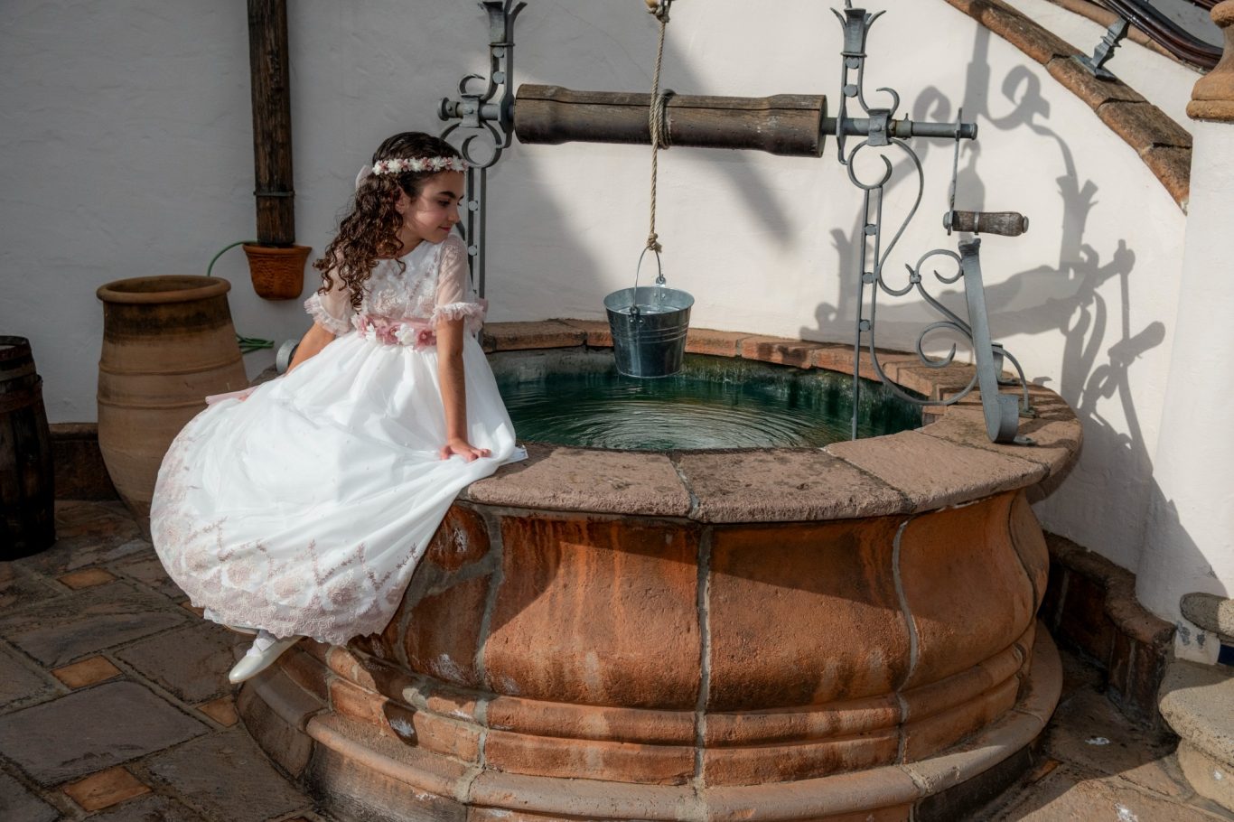 Comuniones en Capricolor Niña con vestido blanco sentada en el borde de un pozo decorativo. - Capricolor