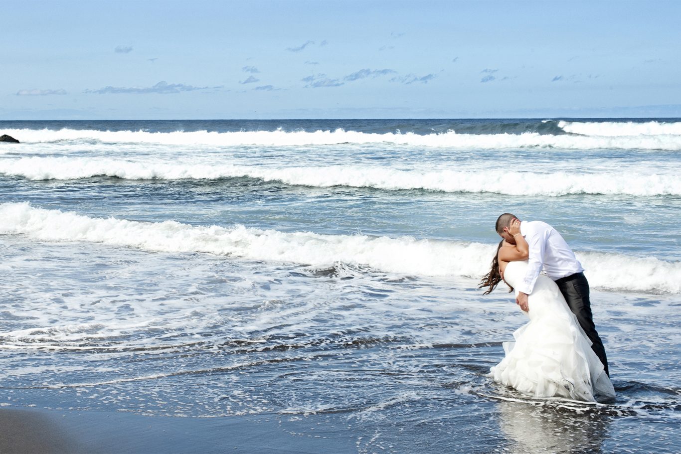 Reportaje de boda en Exterior. Pareja de novios besándose en la playa, con olas y cielo despejado de fondo.- Capricolor