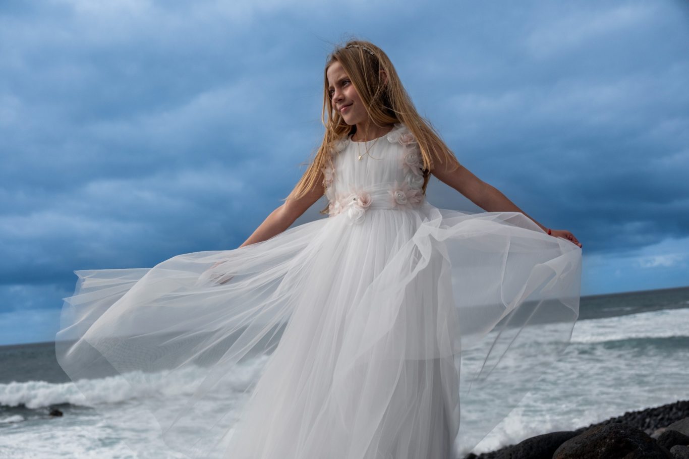 La Primera Comunión de tu hijo-a Niña con vestido blanco ondeante, sonriendo frente al mar y bajo un cielo nublado.- Capricolor