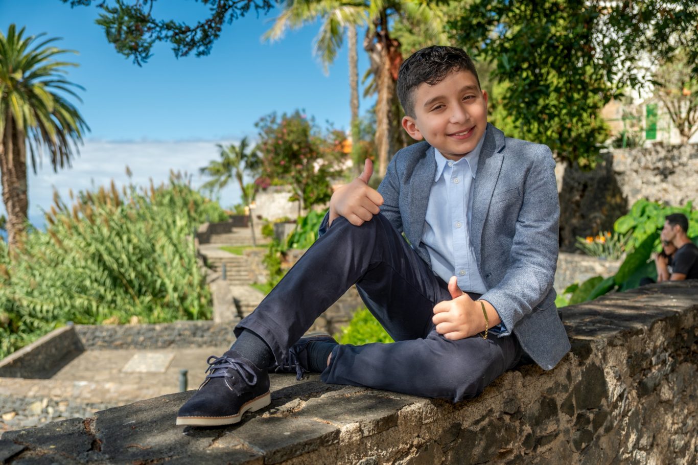 Primeras Comuniones en Capricolor Niño sonriente en un jardín, vestido con traje claro, sentado y haciendo un pulgar arriba.- Capricolor