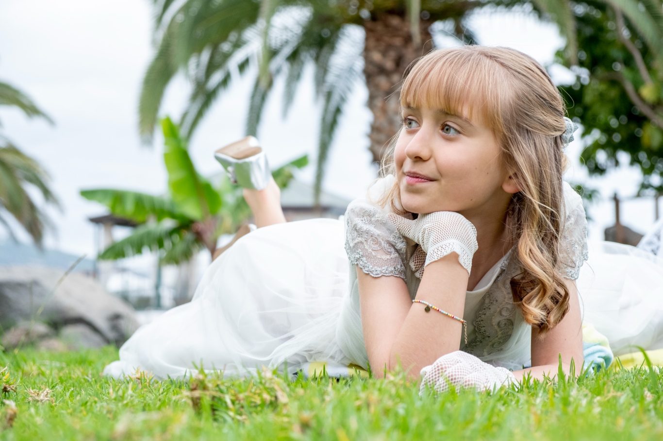 La comunión de tu hijo, una bendición. Niña tumbada en la hierba con un vestido blanco, sonriendo y pensando, rodeada de palmeras.- Capricolor