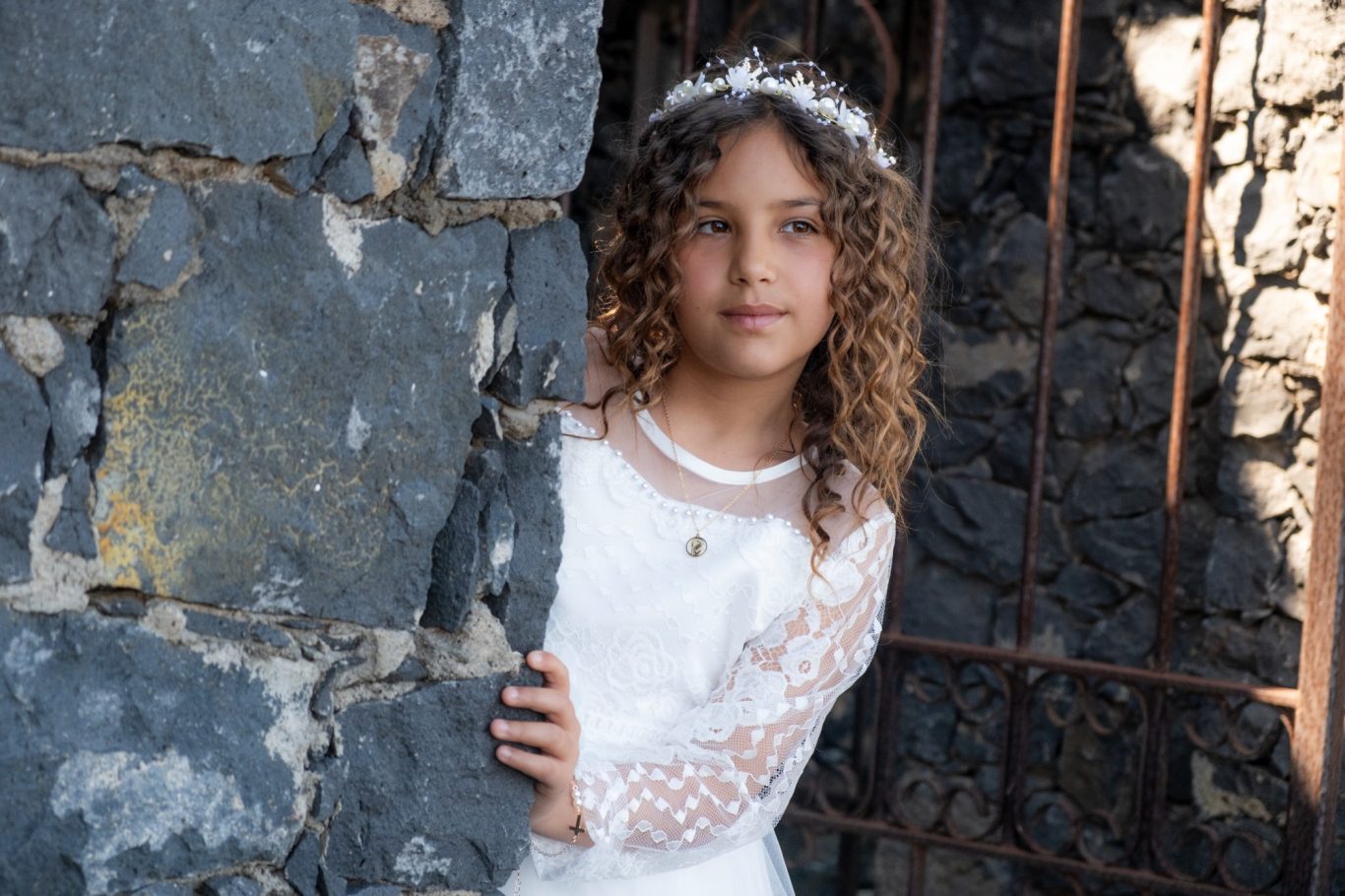 Tu Primera comunión en Capricolor Niña con cabello rizado y vestido blanco, posando junto a una pared de piedra.- Capricolor