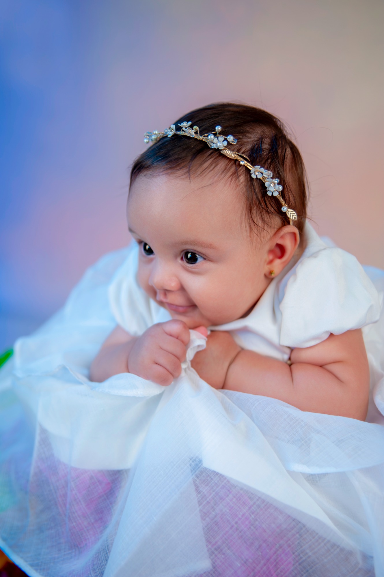 Estudios de Bebés. Bebé con vestido blanco y diadema, sonriendo sobre una superficie colorida.- Capricolor